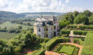Rokokoschloss mit pastellfarbener Fassade, umgeben von symmetrischem Schlossgarten und grüner Landschaft bei Sonnenschein