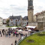 Lebendige Kultur im Herzen Weimars: Open-Air-Veranstaltung vor dem Stadtschloss Mehrere Menschen versammeln sich tagsüber auf einem Platz vor dem Weimarer Stadtschloss zu einer Open-Air-Veranstaltung. Es gibt eine kleine Bühne mit weißen Sonnenschirmen und Sitzgelegenheiten. Im Hintergrund sind der Bastille-Turm, historische Gebäude und Teile eines Gartens mit Blumenbeeten zu sehen.