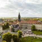 Das Weimarer Stadtschloss mit Open-Air Veranstaltung (von oben) Luftaufnahme des Weimarer Stadtschlosses mit Bastille-Turm bei Abendlicht. Vor dem Schloss findet eine Veranstaltung mit vielen Menschen, Sitzplätzen und weißen Pavillons statt. Im Vordergrund sitzen Besucher auf Picknickdecken im Schlossgarten. Im Hintergrund sind die Stadt Weimar mit Häusern, Kirchtürmen und Bäumen zu sehen.